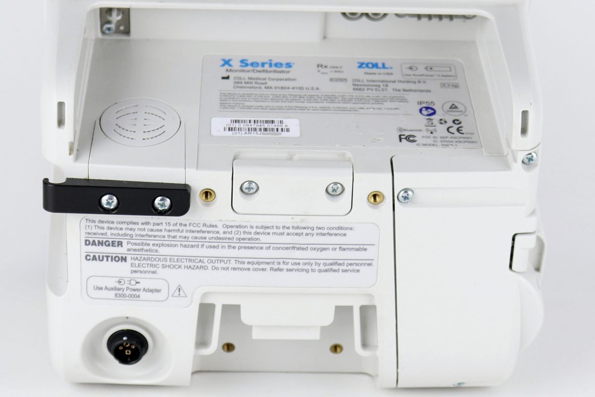 A close view of the back of the monitor showing captive screws, a hinged access panel, the moulded identification plate, and printed hazard warnings.