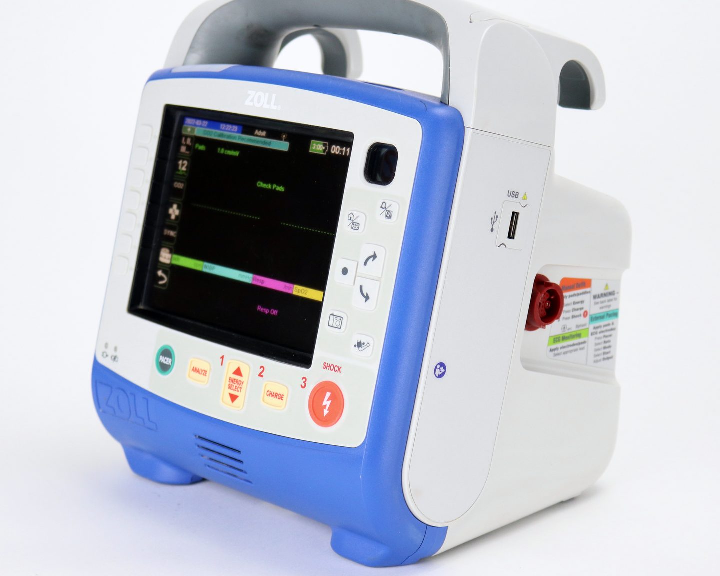 A ZOLL X Series monitor/defibrillator seen from the front left, its carry handle up and its display on, standing on a plain studio surface.